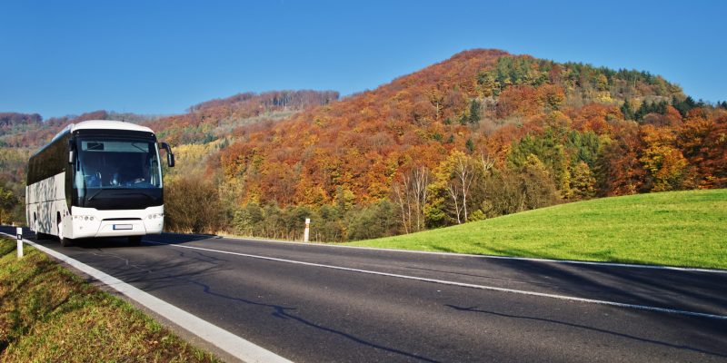 White Bus arriving at the asphalt road through the valley below the wooded mountain of glowing autumn colors. Clear sunny day with blue skies.